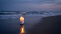 Glowing candle on wet sand reflects light as ocean waves roll onto shore during peaceful twilight evening creating serene coastal atmosphere for meditation.
