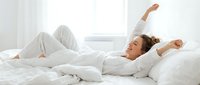 Woman with brown hair and light skin tone stretching in bed after waking, wearing white pajamas. Suitable for wellness, lifestyle, and morning routine content.