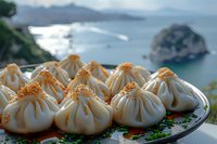Steamed dumplings with sesame seeds served on decorative plate against scenic ocean coastline background, traditional Asian cuisine close-up photography.