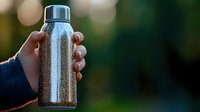 Hand holding stainless steel water bottle with condensation droplets outdoors for hydration and eco-friendly lifestyle.