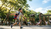 Young Asian boy in striped shirt running joyfully across wooden playground bridge with climbing equipment and green trees in sunny park setting.