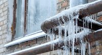 Frozen water pipes with dangerous icicles hanging from residential building gutter system during harsh winter weather conditions causing potential property damage.