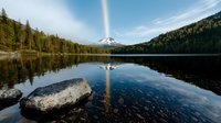 Tranquil mountain lake with snow-capped peak reflection in calm waters, surrounded by evergreen forest. Large boulder in foreground creates dramatic composition.