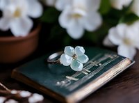 Delicate silver flower ring with pale blue gemstone center resting on vintage green book with white blooms in soft focus background for jewelry photography.