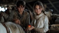 Young couple in vintage clothing at rustic barn stable, woman holding red apple, atmospheric lighting creates moody medieval or historical scene with horses.