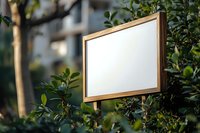 Wooden frame mockup surrounded by green leaves in natural outdoor setting. Empty white display board against blurred garden background for advertising.