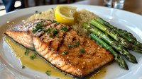 Grilled salmon fillet with butter sauce, fresh herbs, served with quinoa and roasted asparagus spears on white plate, garnished with lemon slice.