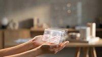 Hands holding transparent glass cube with holographic digital interface display in modern office workspace environment.