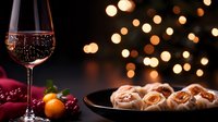 Glass of sparkling wine with cinnamon rolls and tangerines on dark background with festive bokeh lights, cozy winter holiday atmosphere.