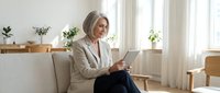 Professional mature blonde businesswoman using tablet computer while sitting on modern sofa in bright office interior with natural lighting.