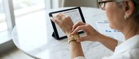 Professional businesswoman with glasses using tablet computer at modern office desk reviewing financial data and analytics charts for business planning.