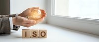 Businessman hands protecting glowing light bulb over ISO wooden blocks representing quality management system standards and certification processes.