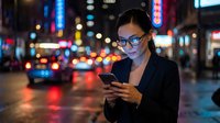 Young Asian woman wearing glasses and dark blazer using smartphone on neon-lit urban street at night with blurred city lights and traffic in background.