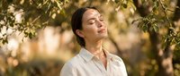 Young Asian woman in white shirt enjoying peaceful moment under tree branches with eyes closed, expressing serenity and mindfulness in natural outdoor setting.