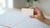 Adult male hand holding blank white card against neutral interior background with natural light, suitable for business branding and marketing mockups.