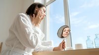 Young woman with dark hair and fair complexion examining her face in handheld mirror near window, wearing white shirt. Skincare routine, beauty reflection, morning grooming, cosmetic assessment.