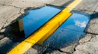 Puddle reflecting blue sky with yellow road marking line on cracked asphalt pavement, suitable for urban street photography and weather-related design projects.