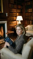 Middle-aged Caucasian woman in professional business attire reading leather-bound book in luxurious library study. Warm fireplace glow, mahogany shelves, classic lamp lighting elegant home office interior.