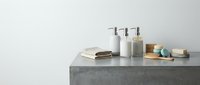 Modern minimalist bathroom counter with concrete surface featuring white pump bottles, natural soap bars, wooden brush and spa accessories for wellness routine.