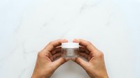 Male hands holding clear cosmetic jar with white cap against marble background for skincare product display.