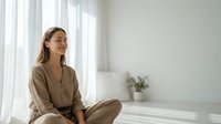 Young Caucasian woman in beige linen outfit sitting peacefully by window in minimalist white bedroom practicing mindfulness meditation for wellness and relaxation.
