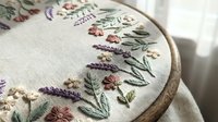 Delicate hand embroidered floral design with lavender and wildflowers on natural linen fabric stretched in wooden hoop for needlework and crafting projects.