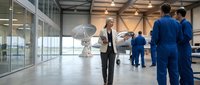 Woman in business attire holding umbrella indoors with team in blue uniforms in modern warehouse or industrial facility space.