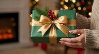 Woman holding green Christmas gift box with gold ribbon bow and holly decoration in cozy living room with fireplace and warm lights during holiday season.