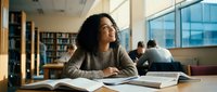 Young African American woman studying in modern university library with books and laptop, focused on academic research and learning goals.