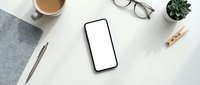 Smartphone with blank screen surrounded by coffee cup, eyeglasses, notebook, pen, succulent plant and wooden clip on white desk workspace.
