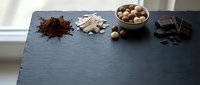 Dark slate board with cocoa powder, white chocolate pieces, hazelnuts in bowl and dark chocolate chunks arranged for baking preparation.