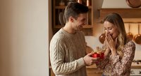 Young couple sharing romantic moment as man presents gift box to surprised woman in cozy kitchen setting during holiday celebration.