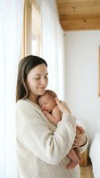 Young mother in cream sweater tenderly holding sleeping newborn baby in bright modern home with wooden accents and natural light streaming through windows.