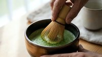 Mature man with medium skin tone whisking matcha powder into hot water with traditional bamboo brush in black ceramic bowl, preparing ceremonial green tea beverage for culinary and wellness content.