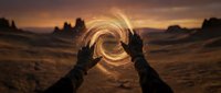Hands creating magical light spiral in desert landscape at sunset with dramatic orange sky and rock formations in background.