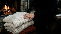 Cozy fireplace setting with person holding stack of knitted sweaters and blankets, warm interior design for home comfort and relaxation.