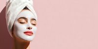 Woman with white facial mask and towel wrapped around head enjoying skincare routine against soft pink background with shadow.