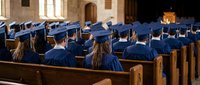 Graduates in blue caps and gowns seated in wooden pews during university commencement ceremony in historic chapel with warm lighting and stone architecture.