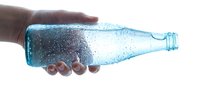 Person with dark skin tone holding a chilled glass bottle filled with sparkling blue carbonated beverage covered in condensation droplets against white background.