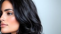 Young Hispanic woman with flawless skin, glossy black wavy hair, and natural makeup looking sideways against light gray background, beauty portrait closeup.