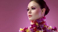 Young Caucasian woman with elegant makeup and updo hairstyle adorned with vibrant purple and yellow orchids around neck on pink background, fashion beauty portrait.