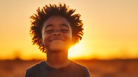 Young African American boy with curly afro hair smiling with eyes closed against golden sunset background, radiating joy and innocence in warm sunlight.