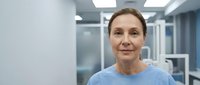 Middle-aged Caucasian female healthcare professional in blue scrubs smiling confidently in modern medical facility corridor with bright lighting.