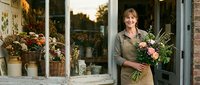 Mature woman florist holding fresh flower bouquet outside rustic flower shop storefront with blooming plants and greenery displayed in windows for retail business.