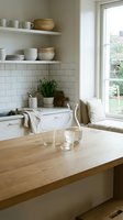 Bright, minimalist kitchen interior with wooden countertop, white subway tile backsplash, open shelving with ceramics, potted herbs, and natural light from large window. Suitable for home decor, interior design, and lifestyle publications.