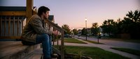 Young man sitting on wooden steps during sunset in suburban neighborhood, contemplating life while looking at peaceful residential street.