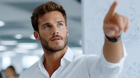 Young professional businessman pointing at whiteboard during office presentation or meeting with colleagues in modern workplace.