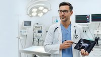 Confident male doctor with glasses holding tablet in modern operating room with surgical lights and medical equipment for healthcare technology.