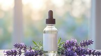 Glass dropper bottle with clear serum surrounded by fresh lavender flowers on white surface. Natural skincare product for aromatherapy and wellness routines.