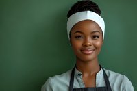 Young African American female chef wearing white headband and gray apron smiling confidently against green background, representing culinary professionalism.